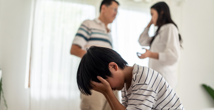 A child is upset, sitting on a couch, and holding their head in their hands. Two adults are talking in the background.