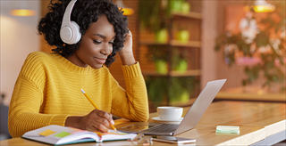 Woman wearing headphones, looking at her computer and writing in a book