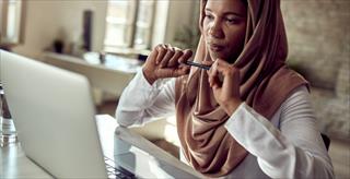 Pensive Muslim businesswoman working on laptop at her office desk.