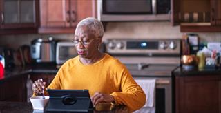 A senior African American woman sits at her kitchen counter and works on her digital tablet that has a keyboard connected and is resting on a stand.